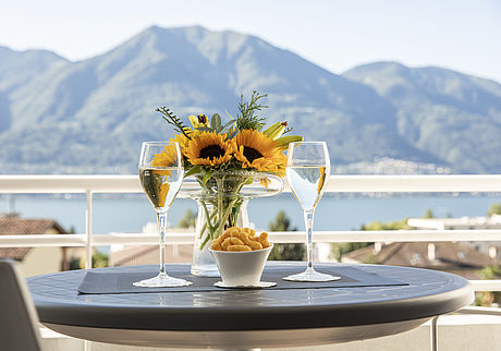Zwei volle Prosecco-Gläser und eine Vase mit Sonnenblumen auf einem Tisch mit Blick auf den See und eine Berglandschaft